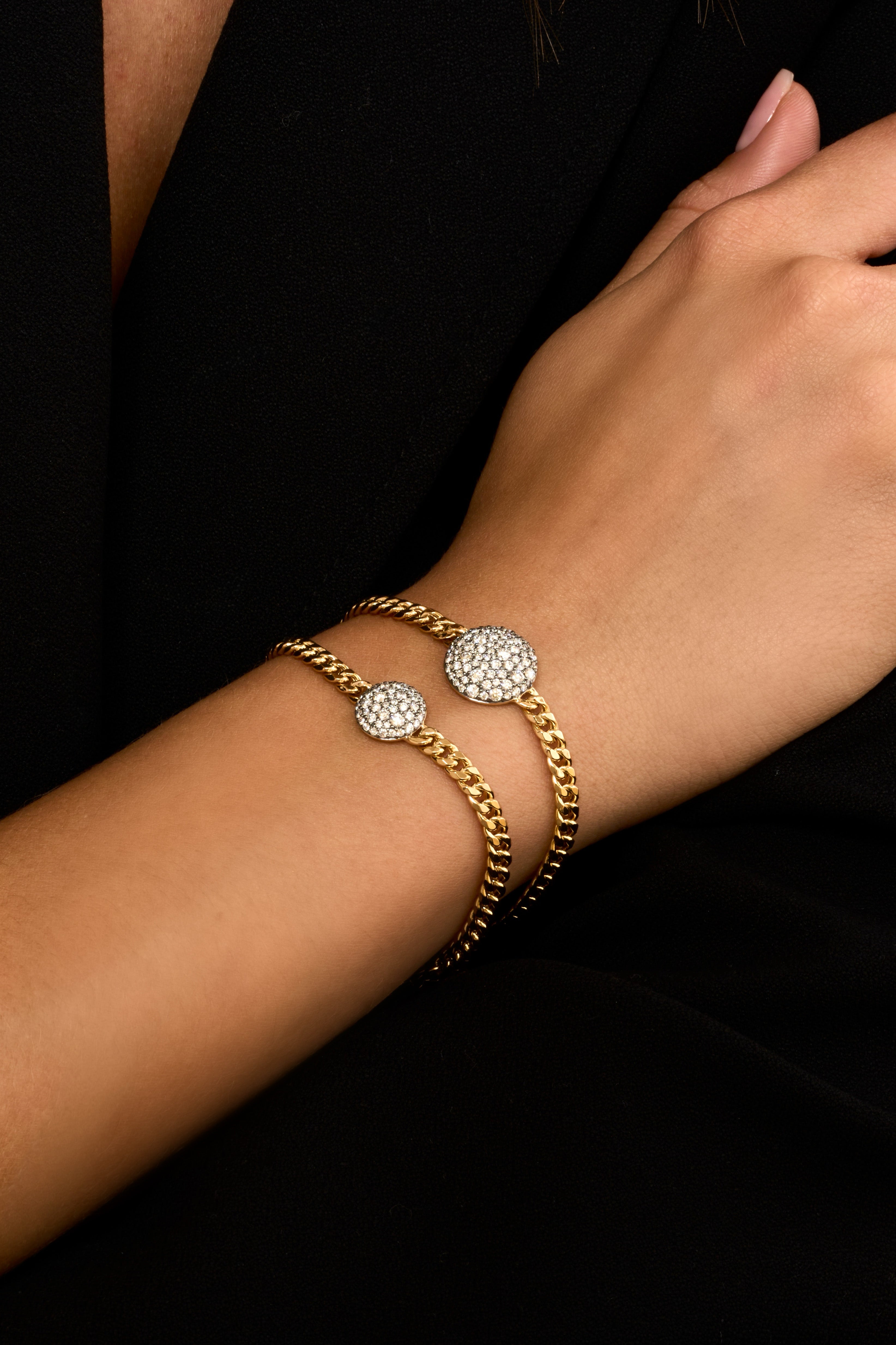 A close-up of a person's wrist wearing Diamond Prima Bracelets in 18k yellow gold with a curb link chain and round colorless diamond pave accent set in black rhodium.